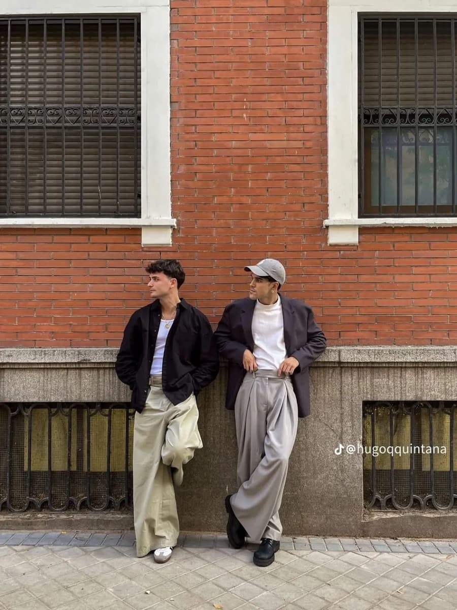 Two men against a brick wall in overshirts and wide pleated trousers